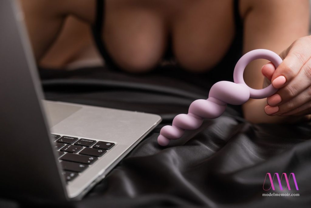 Woman holding lilac anal beads next to laptop while lying on bed.